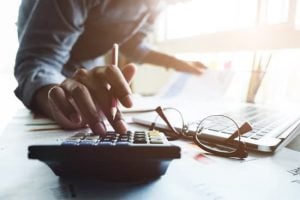 Close up of businessman or accountant hand holding pen working on calculator to calculate business data, accountancy document and laptop computer at office, business concept