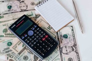 A calculator, pen, and small notebook on top of dollar bills