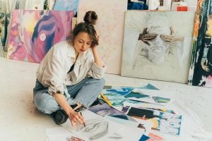 Woman sitting on the floor while sketching and surrounded by various paintings