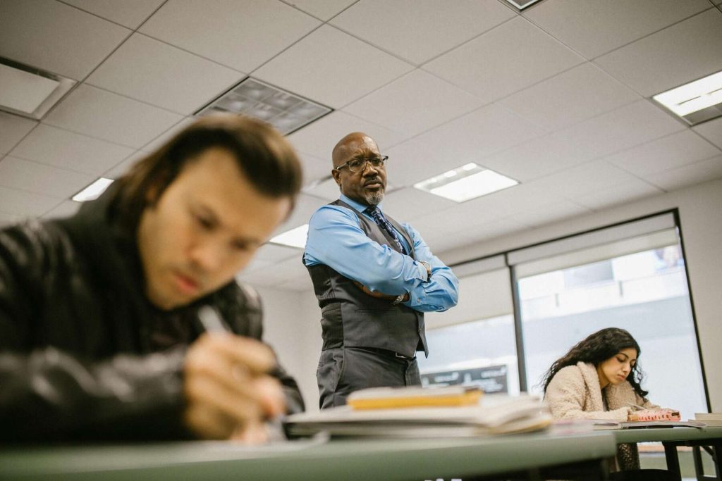 A teacher wearing a blue long sleeve with a vest and tie proctors his students during an examination