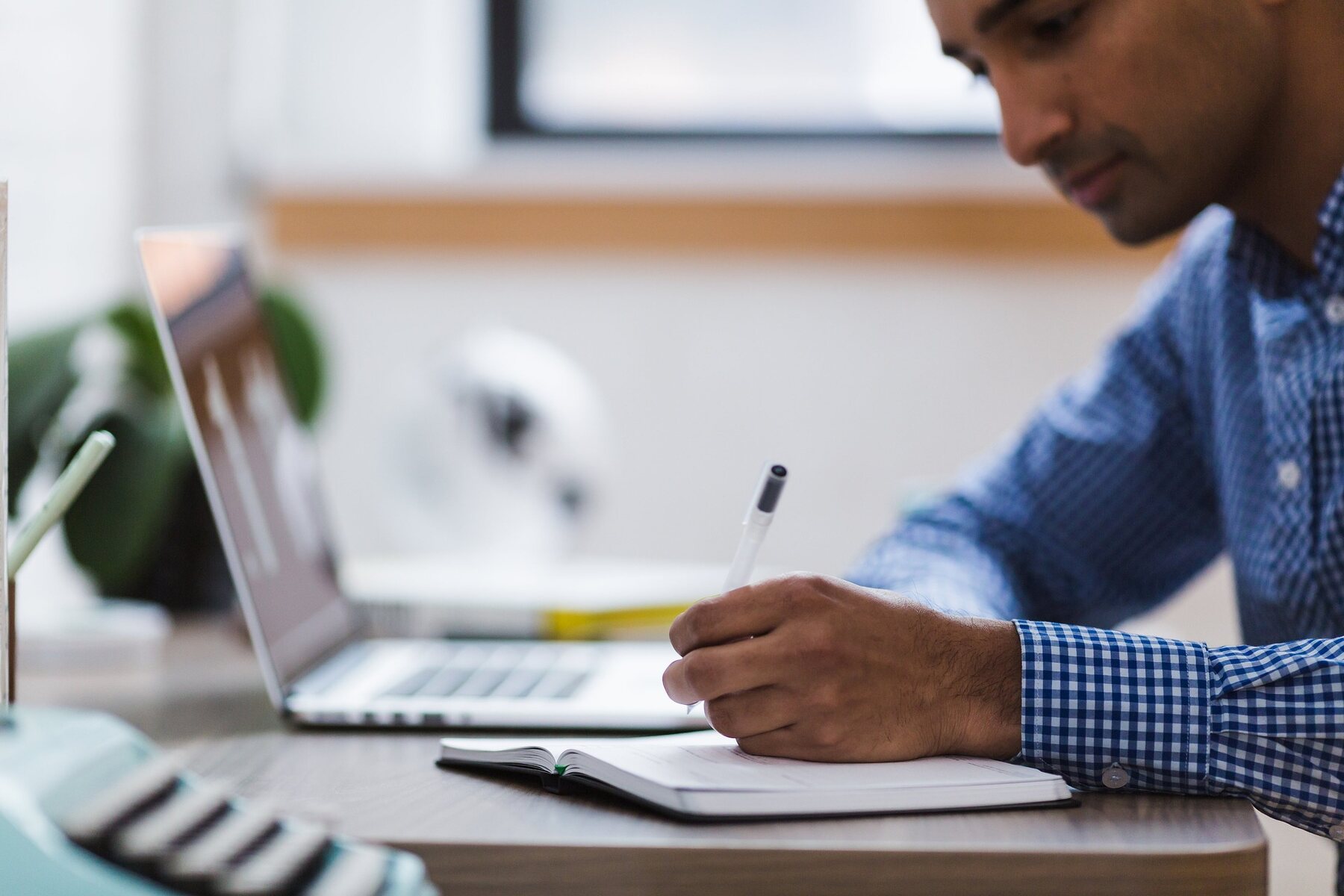 A man writing on a notebook at a desk with a laptop nearby