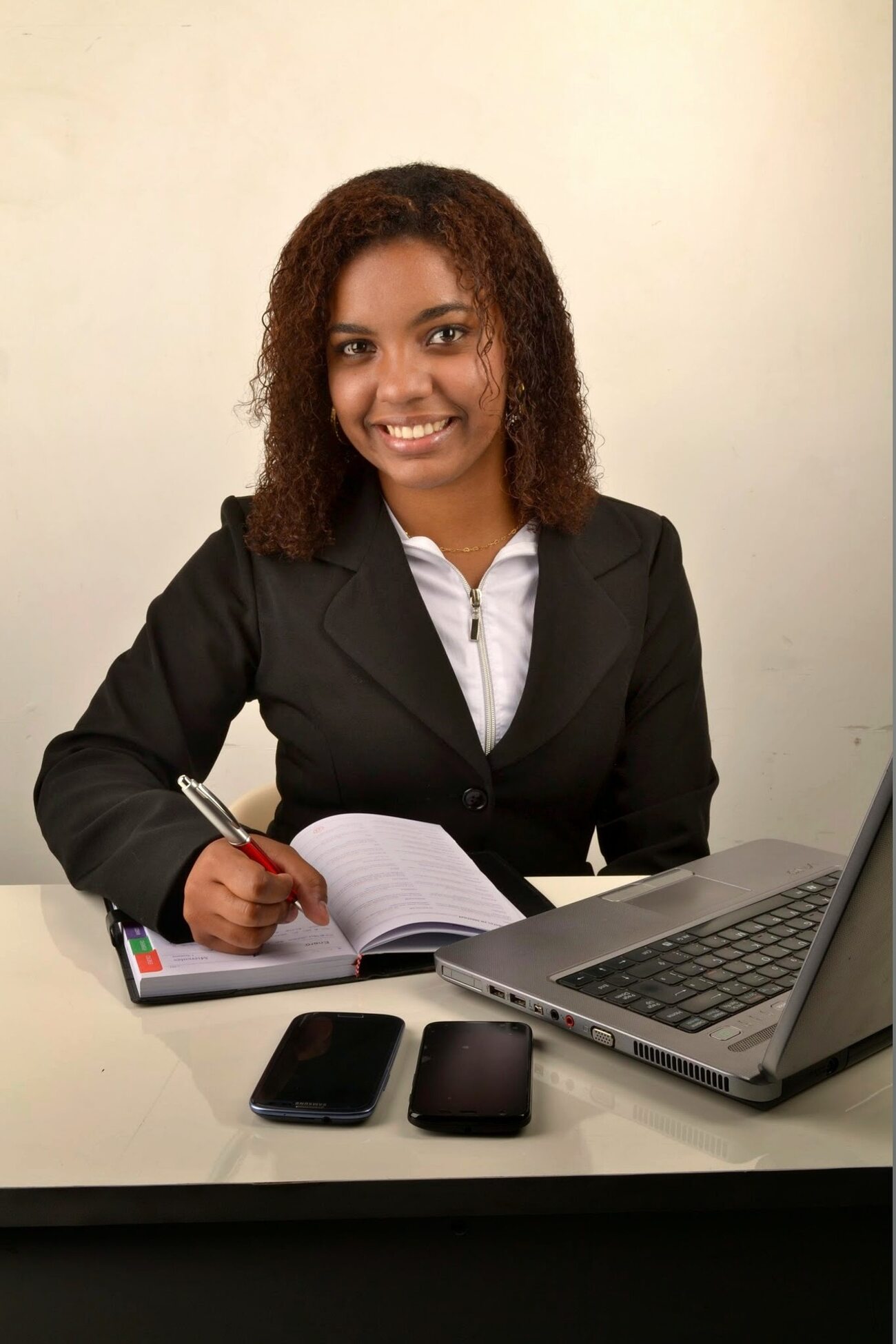 Woman in business attire writing notes on a her planner with her laptop and phones on the table