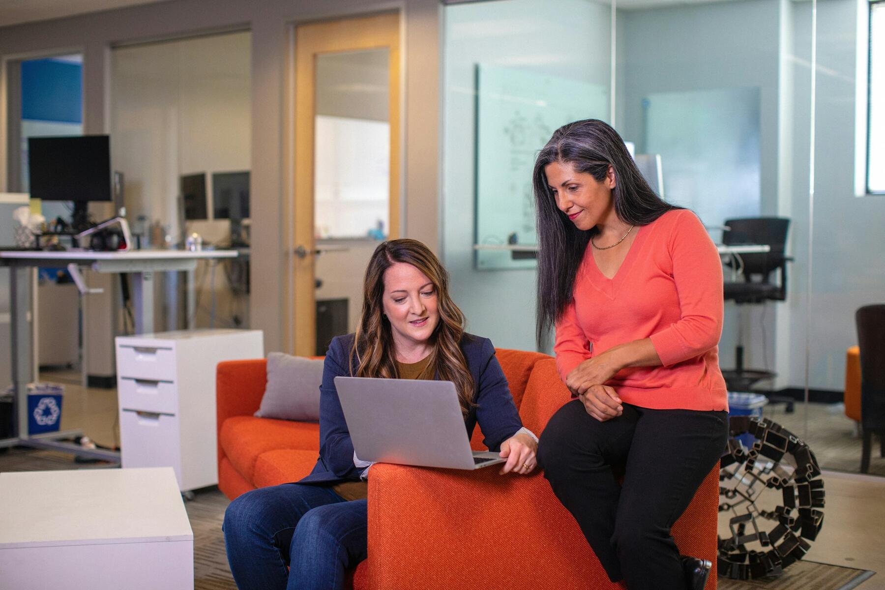 Two women working on a laptop while sitting on an orange couch
