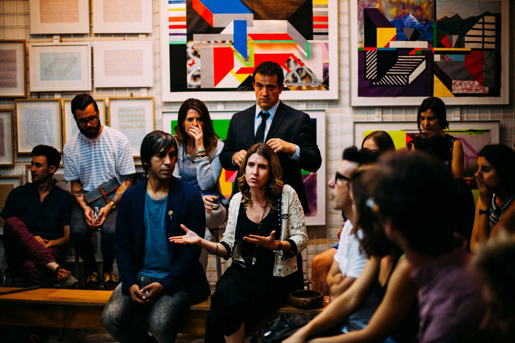 A diverse group of people sitting on benches in front of a painting at an art gallery discussing some matters