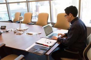 Man sitting on a long conference table while working on his laptop