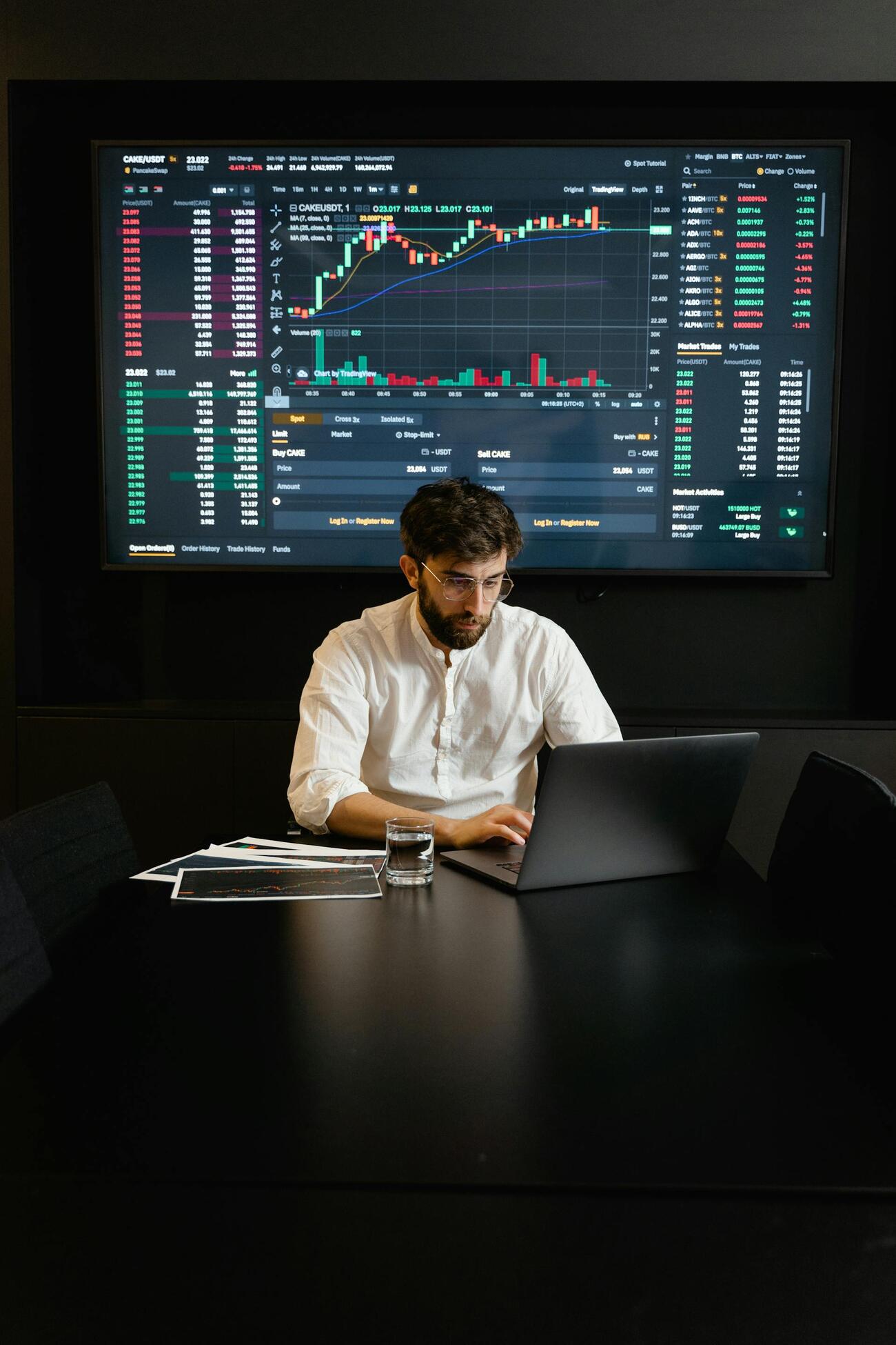 A man at a table with a laptop, with stock market data on the screen behind him